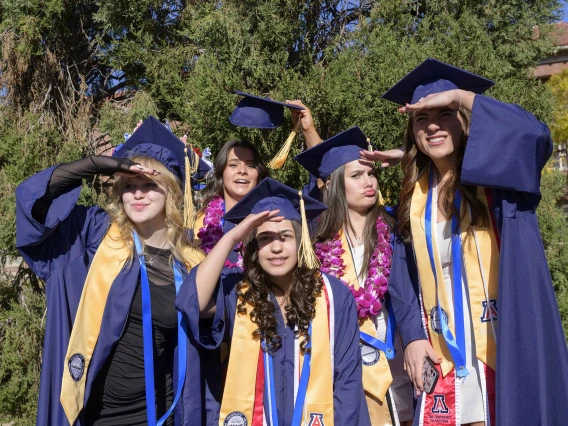 Five University of Arizona College of Nursing graduates in caps and gowns stand together, shielding their eyes from the sun. 