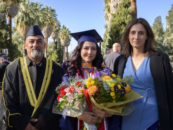 A young woman dressed in a graduation cap and gown holds flowers as she is flanked by her parents. 