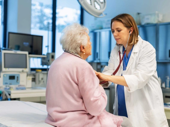 A doctor examines a patient wearing a pink robe sitting on the edge of a bed