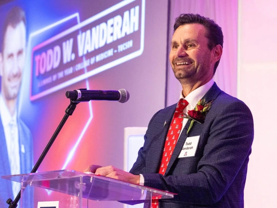 Dark haired man in suit and tie standing at a podium smiling. 