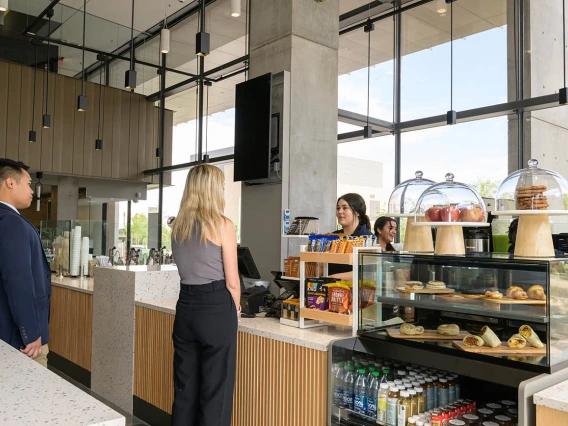 A woman with long blonde hair stands at a coffee bar counter with pastries in a case. 