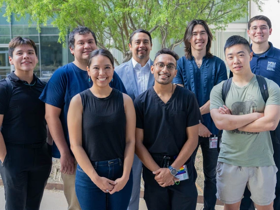 Eight people stand in a group in an outside setting all smiling. 