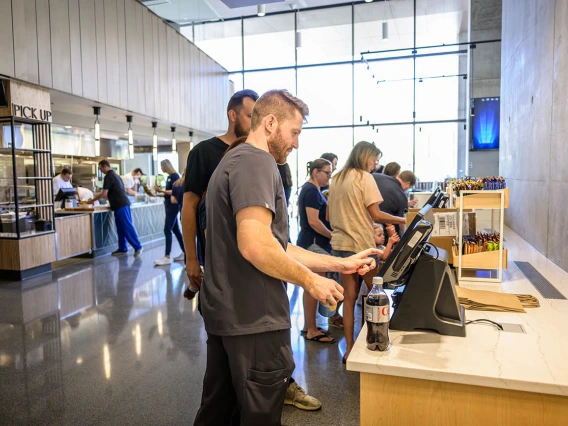 Several people stand at computer screens in a cafe setting placing their orders. 