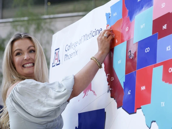 A light skinned woman with blonde hair smiles big as she puts a pin in a big map of the U.S. 