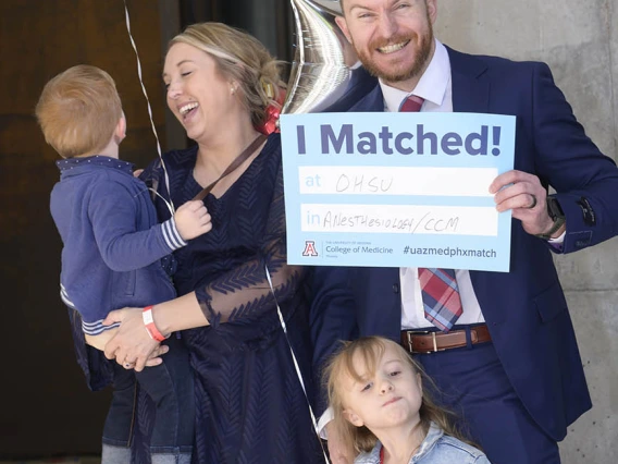 A family stands together as the dad holds an "I Matched" sign and his wife looks at the boy she is holding and smiles. Young girl standing in front of them.