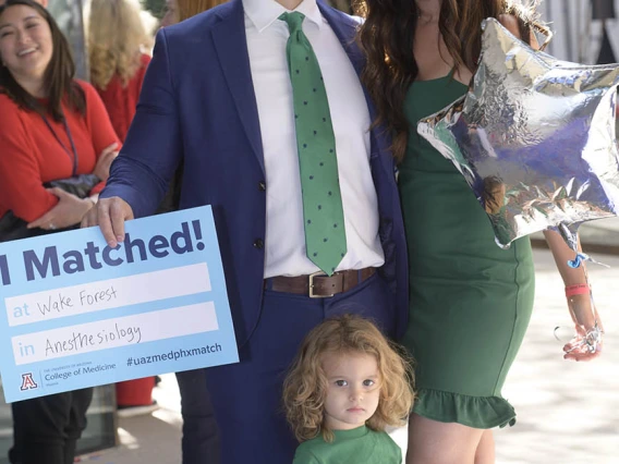 A light skinned man in a suite with his wife and child smile. He holds a poster that says "I Matched"