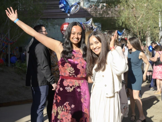 Two young woman stand side by side smiling and waiving. Both have long dark hair and light brown skin. 