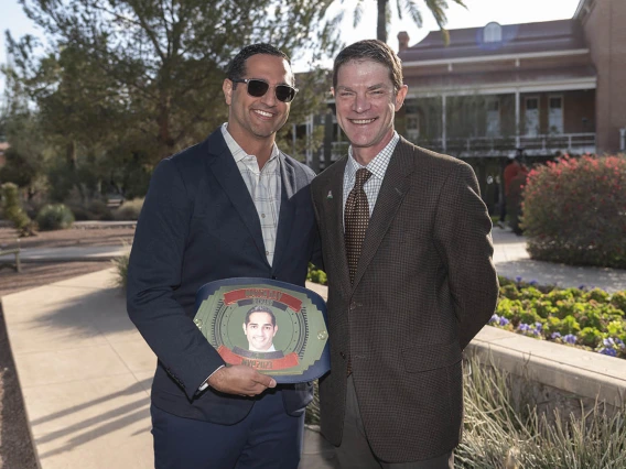 Two men in suites stand outside smiling. The younger man with dark hair holds an oval plackard with his photo on it. 