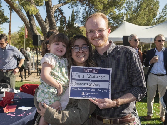 A young couple holding a little girl smile while holding a Match Day sign. All three are light skinned.