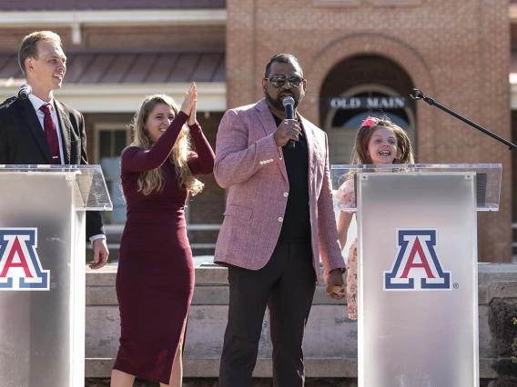 A man in a light pink jacket speaks into a microphone while holding the hand of a young girls who is smiling. 