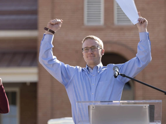 A smiling young adult man with light skin and hair in a dress shirt holds up both arms in celebration. 