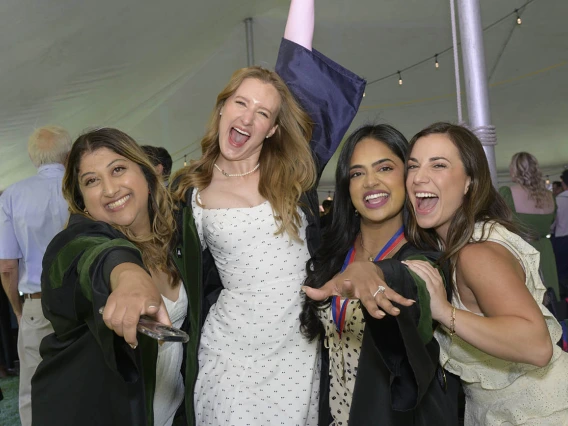 Four young women in graduation regalia smile and hug in celebration. 