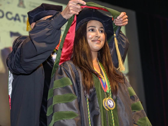 A young woman with long dark hair wearing a cap and gown has a sash placed over her shoulders. 