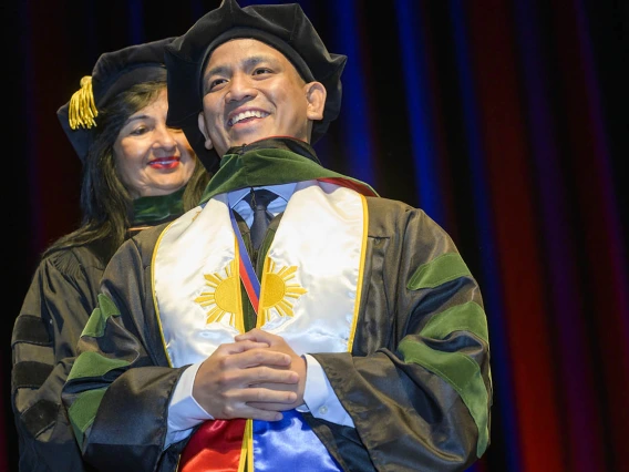 A young man in graduation regalia with a white stohl around his neck, smiles as a professor places a sash over his shoulders. 