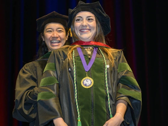 A young woman with long dark hair wearing a graduation cap and gown smiles as a faculty member behind her smiles as well. 