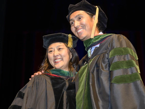 A tall young man in graduation regalia side hugs a professor, also in graduation regalia, as both smile. 