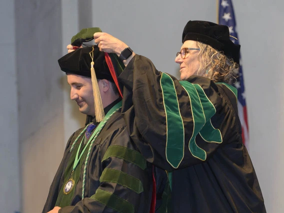 Side view of a young man in graduation regalia has a sash placed over his head by a female professor, alos in graduation regalia.