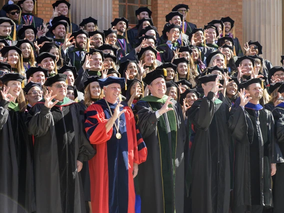 Dozens of graduates and faculty in graduation regalia stand on stairs making the Arizona Wildcat sing with their fingers. 