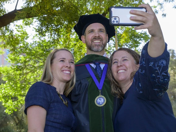 Two women stand on each side of a tall man in graduation cap and gown as one of the women takes a selfie. 