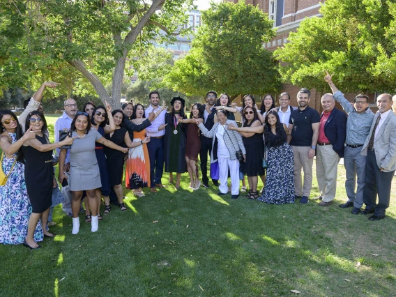 Around 25 family members stand on both sides of a medical shool graduate wearing a cap and gown as they all gesture towards the graduate. 