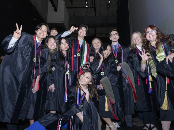 About a dozen pharmacy graduates in caps and gowns bunch together smiling and cheering.