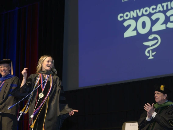 A young white woman in graduation gown bounds across the stage in front of a large screen that reads "Convocation 2023"