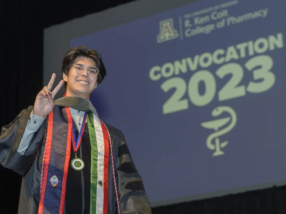 A young man in a graduation gown flashes a peace sign and smiles as he crosses the stage. 