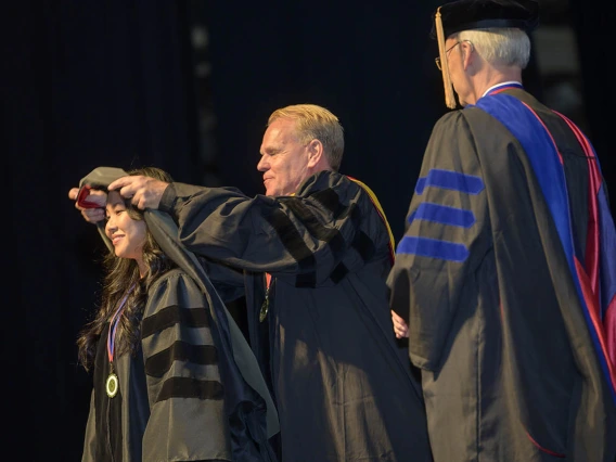 A young Asian woman in a graduation gown has a sash placed over her shoulders by a middle-aged white professor in a black graduation gown. 