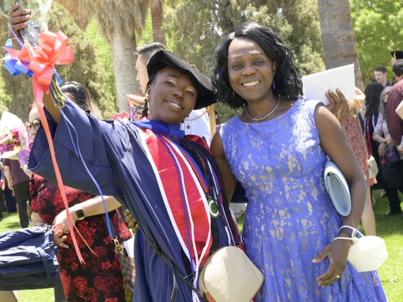 Two young Black women, one in a dress and the other in a graduation cap and gown, side-hug and smile.