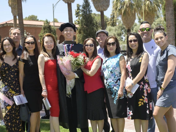 A young male graduate in cap and gown smiles while holding flowers as he stands with about 10 family members outside.