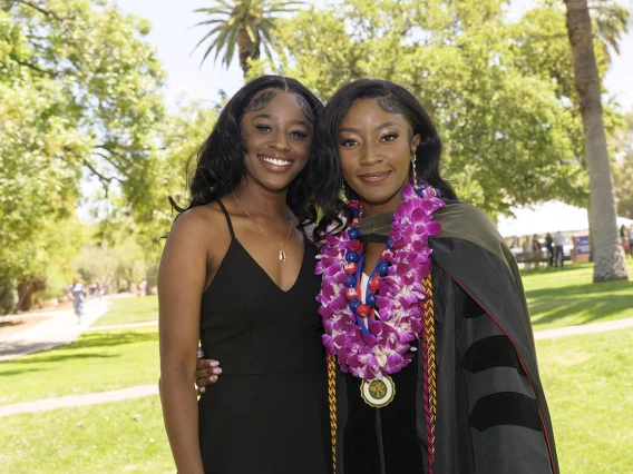 Two young Black women, one in a dress and the other in a graduation gown, side-hug and smile.