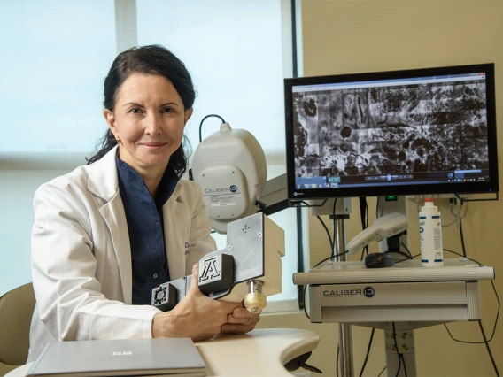 Clara Curiel Lewandrowski, MD, in front of a screen showing the images produced by a confocal microscope. She holds the smaller, portable version she is developing for wider use by doctors.