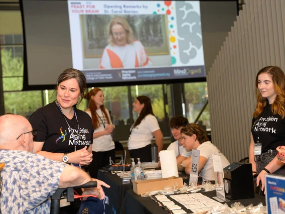 Two women wearing t-shirts that read "Precision Aging Network" talk with an older man in a wheelchair inside a large open room with other people in the background. 