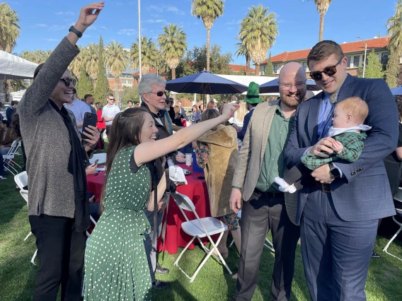 A light-skinned man in a suite holds a baby while several people are trying to get the baby to look their way for a photo.