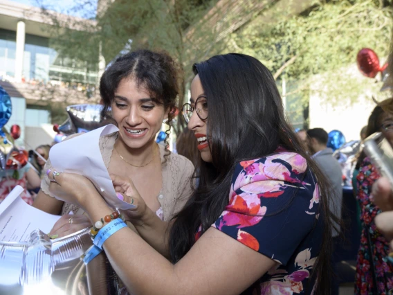 Two young adult women with dark hair look at each other's letters and smile. 