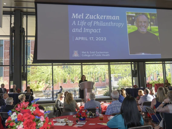 Crowd of people sitting at tables watching Dr. Iman Hakim, dean of the University of Arizon Mel and Enid Zuckerman College of Public Health, speak at a podium.