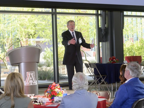 An older light-skinned man in a suite stands on a stage talking to a crowd. 