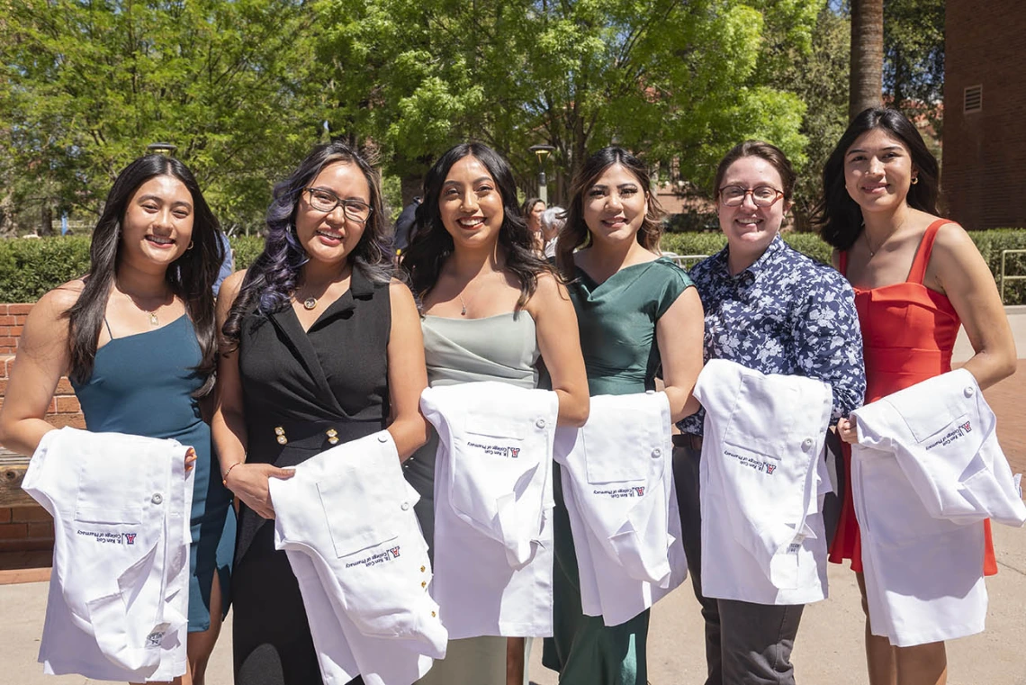 Six young women stand side-by-side outside with white coats draped over their arms. 