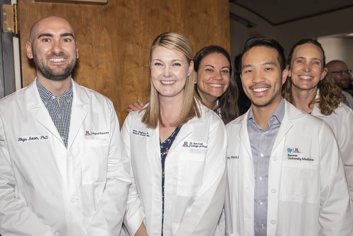 Five men and women Pharmacy faculty members wearing white coats standing side-by-side smiling. 