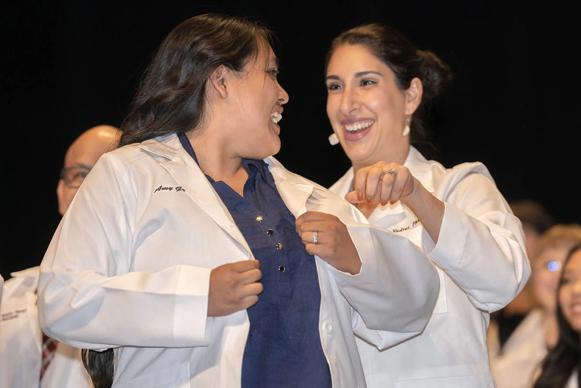 Two women wearing Pharmacy white coats smile as the younger woman puts the coat on. 