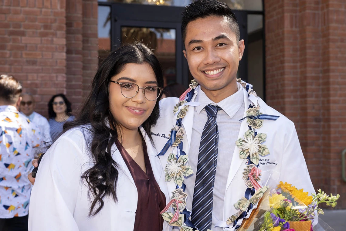 A yong woman and man wearing pharmacy white coats stand next to each other smiling. 