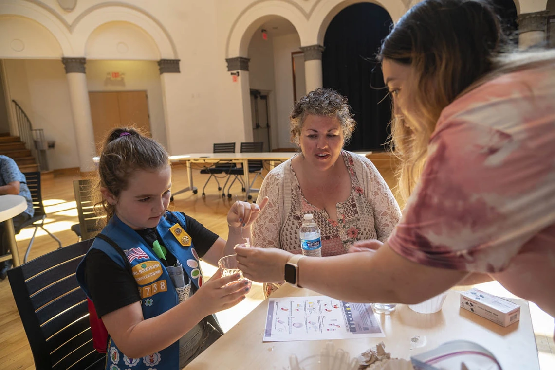 A young girl scout works with a volunteer at an activity table as her mother watches. 