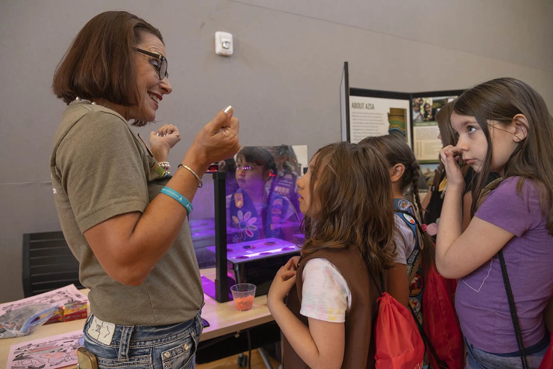 A middle-aged woman with dark brown hair stands next to several girl scouts explaining a display at a table. 