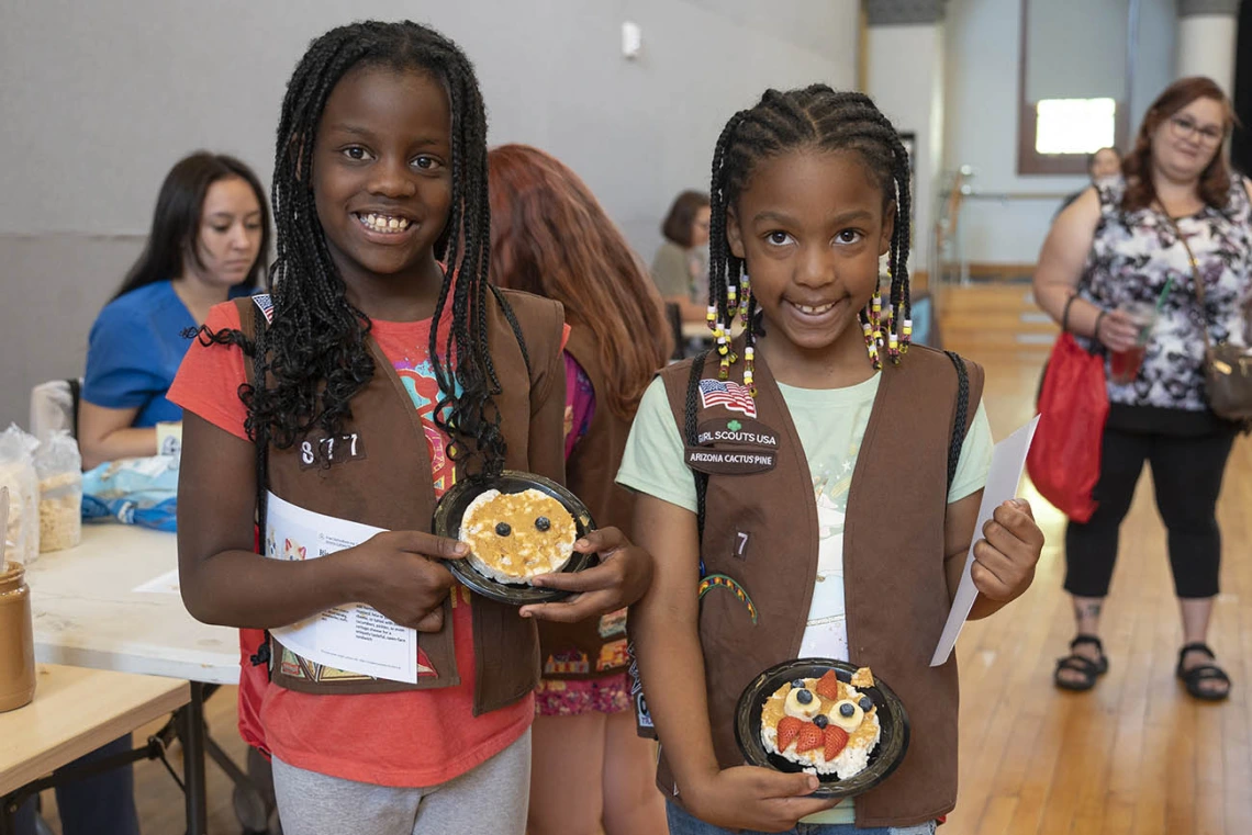 Two brown-skinned young girls in girl scout uniforms smile and hold up decorated rice cakes.