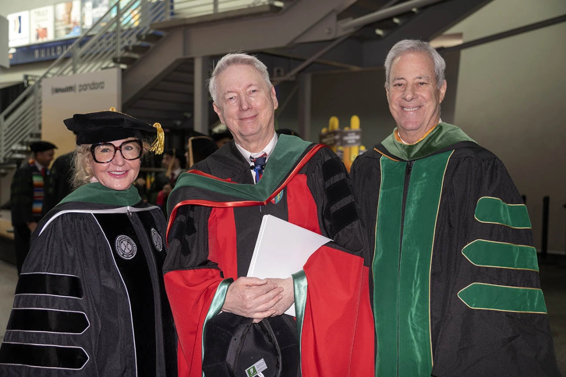 An older woman and two older men, all in graduation regalia, smile as they stand next to each other. 