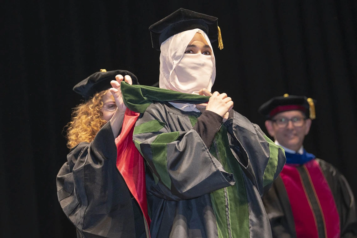 A young woman wearing graduation regalia and a head and face covering is presented with a sash for graduation.