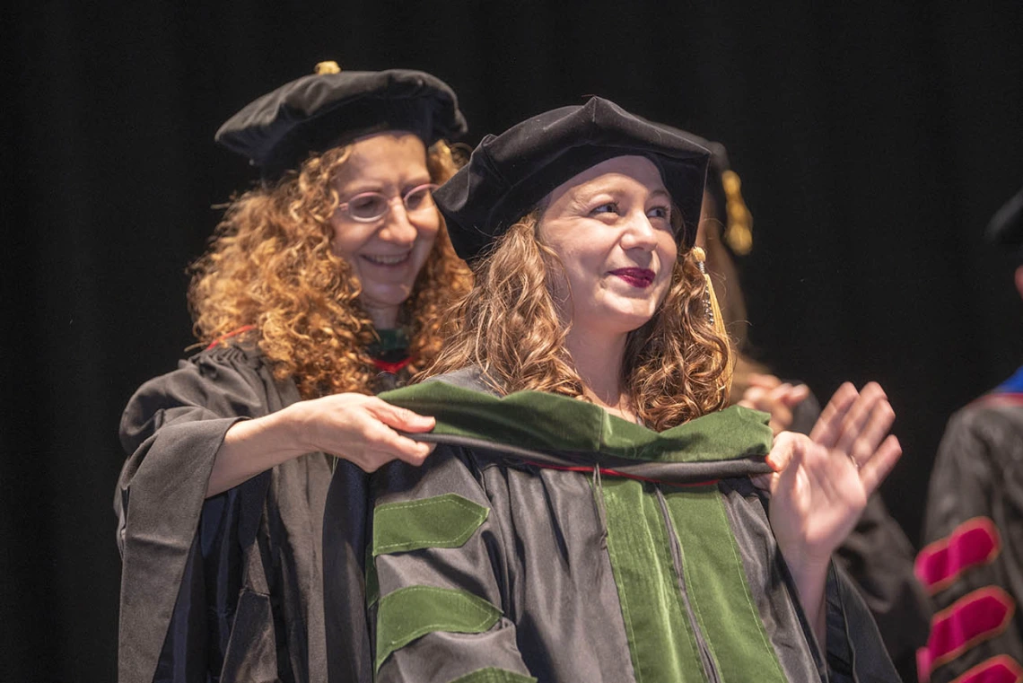 A young woman wearing graduation cap and gown waves as a sash is placed over her shoulders by a faculty member.