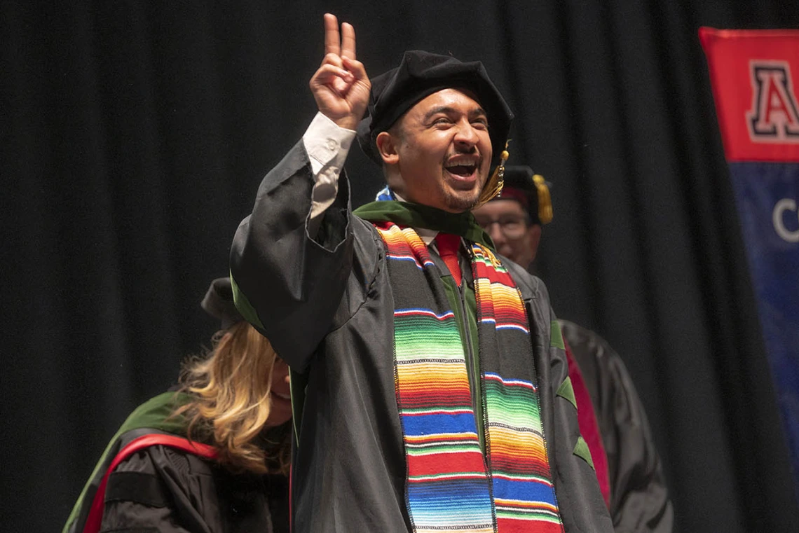 A young man wearing a graduation cap and gown smiles while holding up two fingers.