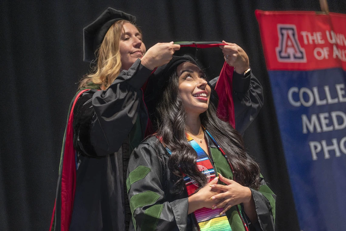 A young woman wearing a graduation cap and gown looks up and smiles as her graduation sash is placed on her shoulders by a faculty member.