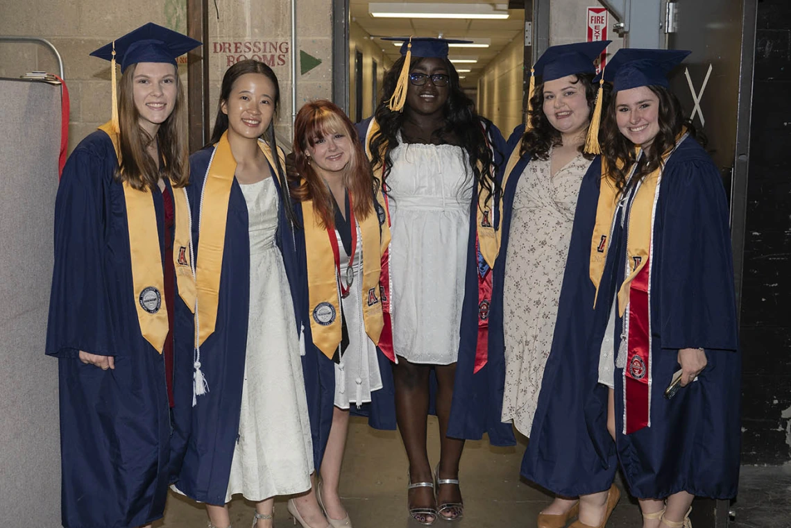 Six female graduates dressed in cap and gown wait backstage in Centennial Hall before receiving their degrees.